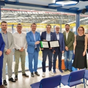 Um grupo de nove pessoas posa em p&eacute; lado a lado para a foto em um ambiente industrial. Ao centro, dois homens seguram e exibem uma placa de reconhecimento. 