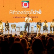 Vis&atilde;o ampla do palco durante evento, com o governador ao centro conduzindo a apresenta&ccedil;&atilde;o e outras pessoas sentadas em cadeiras alinhadas ao fundo. O painel LED exibe &ldquo;Alfabetiza Tch&ecirc; &ndash; Pr&ecirc;mio das Escolas&rdquo;, al&eacute;m de logotipos institucionais do governo estadual e parceiros. A plateia ocupa toda a &aacute;rea frontal do audit&oacute;rio.