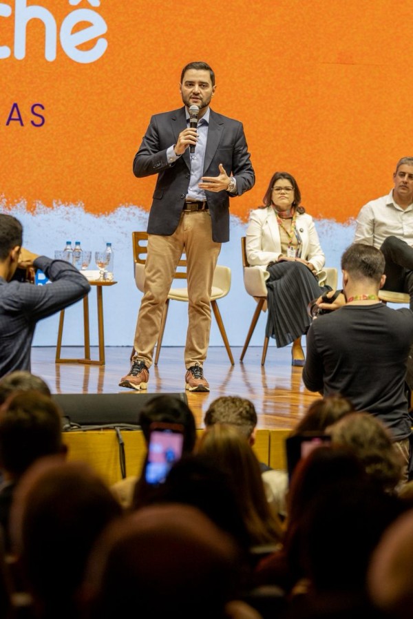 Cena de apresentação no palco, com o vice-governador em pé segurando microfone e gesticulando durante a fala. Atrás, o painel laranja do evento aparece parcialmente, com cadeiras ocupadas por participantes sentados.