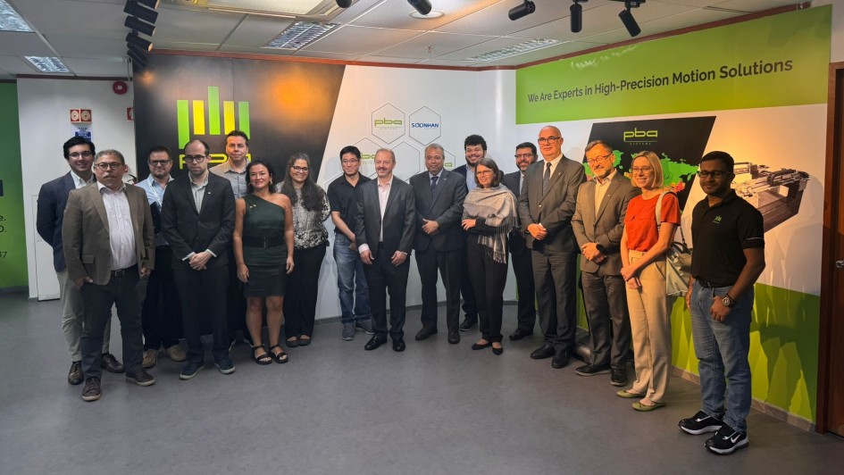 Grupo numeroso reunido em ambiente interno, organizado para registro coletivo diante de um painel institucional. O fundo exibe a identidade visual da empresa, com o texto &ldquo;We Are Experts in High‑Precision Motion Solutions&rdquo;, logotipos corporativos e ilustra&ccedil;&otilde;es t&eacute;cnicas de equipamentos industriais. 