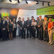 Grupo numeroso reunido em ambiente interno, organizado para registro coletivo diante de um painel institucional. O fundo exibe a identidade visual da empresa, com o texto &ldquo;We Are Experts in High‑Precision Motion Solutions&rdquo;, logotipos corporativos e ilustra&ccedil;&otilde;es t&eacute;cnicas de equipamentos industriais. 