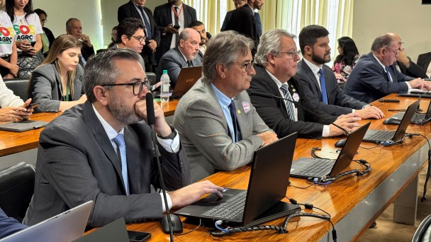 A imagem mostra uma sess&atilde;o institucional em uma sala de reuni&otilde;es ampla, com participantes sentados lado a lado em uma longa mesa de madeira. Cada posi&ccedil;&atilde;o possui notebook aberto, microfone individual, cabos organizados, celulares e garrafas de &aacute;gua. 