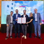Grupo alinhado sobre o palco, posicionado diante de um painel LED com o texto &ldquo;ASSINATURA &ndash; PORTA DE ENTRADA &ndash; FASE 3&rdquo;. Ao centro, um documento aberto &eacute; exibido para registro oficial. 