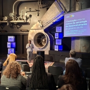 Aqui est&aacute; a descri&ccedil;&atilde;o detalhada da imagem para fins de acessibilidade:

Descri&ccedil;&atilde;o da Imagem
A fotografia captura uma palestra durante um evento preparat&oacute;rio para a Gramado Summit 2026. A cena ocorre em um ambiente com est&eacute;tica industrial moderna e ilumina&ccedil;&atilde;o dram&aacute;tica.

Elementos Principais
A Palestrante: No centro do palco, uma mulher negra veste um terno cinza claro. Ela est&aacute; de p&eacute; sobre um tapete persa, segurando um microfone e gesticulando enquanto fala para a plateia.

O Cen&aacute;rio: O fundo &eacute; composto por uma parede cinza escura com tubula&ccedil;&otilde;es industriais expostas, man&ocirc;metros e uma grande estrutura met&aacute;lica circular que se assemelha a uma turbina ou sistema de ventila&ccedil;&atilde;o.

A Apresenta&ccedil;&atilde;o: &Agrave; direita da palestrante, h&aacute; uma grande tela de proje&ccedil;&atilde;o exibindo um slide com fundo roxo. O texto diz: "A inova&ccedil;&atilde;o est&aacute; no centro da estrat&eacute;gia de desenvolvimento do governo do RS". O slide tamb&eacute;m inclui o desenho de um alvo com um dardo no centro e o logotipo do Governo do Estado do Rio Grande do Sul.

Elementos de Marca: Pr&oacute;ximo &agrave; palestrante, h&aacute; cubos iluminados empilhados exibindo logotipos de parceiros e eventos, como Gramado Summit, Sebrae, Google for Startups e RS Inova&ccedil;&atilde;o.

A Plateia: Em primeiro plano, vemos a parte de tr&aacute;s das cabe&ccedil;as e ombros de v&aacute;rias pessoas sentadas em cadeiras pretas, focadas na apresenta&ccedil;&atilde;o. A ilumina&ccedil;&atilde;o do audit&oacute;rio &eacute; reduzida, destacando apenas o palco e a tela.