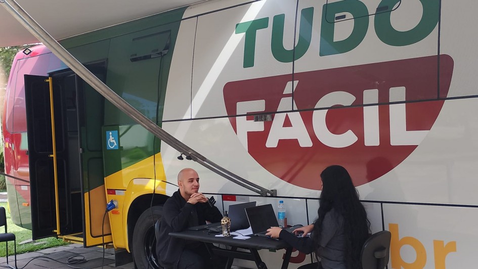 A fotografia mostra a Unidade M&oacute;vel do Tudo F&aacute;cil estacionada em um local p&uacute;blico ao ar livre, em Farroupilha. A cena foca no atendimento ao cidad&atilde;o realizado na parte externa do ve&iacute;culo.

Elementos Principais
O Ve&iacute;culo: &Agrave; esquerda e ao centro, v&ecirc;-se a lateral de um &ocirc;nibus adaptado. Ele &eacute; predominantemente branco com detalhes em verde e amarelo na parte frontal. Na lateral, h&aacute; um grande logotipo com as palavras "TUDO" em verde e "F&Aacute;CIL" em branco dentro de uma forma geom&eacute;trica vermelha. O &ocirc;nibus possui um adesivo com o s&iacute;mbolo internacional de acessibilidade pr&oacute;ximo &agrave; porta.

O Atendimento: Uma mesa preta dobr&aacute;vel est&aacute; montada ao lado do &ocirc;nibus, sob a prote&ccedil;&atilde;o de um toldo branco estendido a partir da lateral do ve&iacute;culo.

Atendente: &Agrave; direita, uma mulher de cabelos longos e pretos, vestida de preto, est&aacute; sentada operando um notebook.

Cidad&atilde;o: &Agrave; esquerda da mesa, um homem jovem, careca e tamb&eacute;m vestido de preto, est&aacute; sentado de frente para a atendente, aguardando ou acompanhando o servi&ccedil;o.

O Ambiente: O ch&atilde;o &eacute; de lajes de pedra cinza irregulares. O local parece ser um parque ou pra&ccedil;a, com gramado vis&iacute;vel ao fundo, &agrave; esquerda. A luz do dia sugere um ambiente externo bem iluminado.

Resumo visual: A imagem ilustra a presta&ccedil;&atilde;o de servi&ccedil;os p&uacute;blicos itinerantes. A estrutura m&oacute;vel do Tudo F&aacute;cil permite que o governo leve atendimento de documenta&ccedil;&atilde;o e servi&ccedil;os digitais diretamente &agrave;s comunidades, como exemplificado pelo atendimento personalizado na mesa externa.