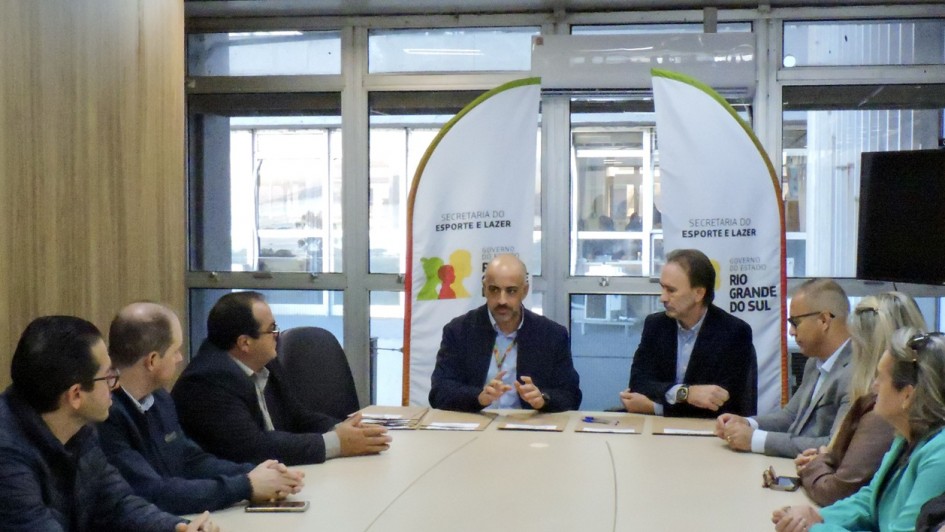 Aqui est&aacute; a descri&ccedil;&atilde;o detalhada da imagem para fins de acessibilidade:

Descri&ccedil;&atilde;o da Imagem
A fotografia registra uma reuni&atilde;o oficial para assinatura de conv&ecirc;nios entre o governo estadual e representantes municipais. A cena ocorre em uma sala de reuni&otilde;es com ilumina&ccedil;&atilde;o natural vinda de grandes janelas ao fundo.

Elementos Principais
A Mesa de Reuni&atilde;o: No centro da imagem, um grupo de pessoas est&aacute; sentado ao redor de uma grande mesa de madeira clara em formato oval.

Ao centro: Um homem calvo, vestindo um palet&oacute; azul-marinho sobre uma camisa clara, fala e gesticula com as m&atilde;os. Ele parece liderar a reuni&atilde;o.

Ao redor: Outros sete participantes (homens e mulheres) est&atilde;o distribu&iacute;dos pelas laterais da mesa. Eles vestem roupas formais e casuais-chiques (palet&oacute;s, blazers e malhas). Alguns olham atentamente para o orador, enquanto outros mant&ecirc;m as m&atilde;os sobre a mesa.

Documenta&ccedil;&atilde;o: Sobre a mesa, em frente a cada participante, h&aacute; pastas ou documentos pardos organizados, prontos para a assinatura dos conv&ecirc;nios mencionados no t&iacute;tulo.

Elementos Visuais de Fundo: Atr&aacute;s do orador central, h&aacute; dois banners verticais (estilo wind banner) brancos com detalhes em verde e laranja. Neles, l&ecirc;-se: "SECRETARIA DO ESPORTE E LAZER" e o logotipo do Governo do Estado do Rio Grande do Sul.

O Ambiente: O fundo &eacute; composto por janelas de vidro com molduras met&aacute;licas que mostram um ambiente externo levemente desfocado. &Agrave; esquerda, h&aacute; um detalhe de uma parede revestida em madeira.

Resumo visual: A imagem captura um momento de articula&ccedil;&atilde;o pol&iacute;tica e administrativa. O foco est&aacute; na formaliza&ccedil;&atilde;o de investimentos para o esporte e lazer, representada pela seriedade dos participantes e pelos materiais de identifica&ccedil;&atilde;o da Secretaria de Estado.