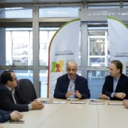 Aqui est&aacute; a descri&ccedil;&atilde;o detalhada da imagem para fins de acessibilidade:

Descri&ccedil;&atilde;o da Imagem
A fotografia registra uma reuni&atilde;o oficial para assinatura de conv&ecirc;nios entre o governo estadual e representantes municipais. A cena ocorre em uma sala de reuni&otilde;es com ilumina&ccedil;&atilde;o natural vinda de grandes janelas ao fundo.

Elementos Principais
A Mesa de Reuni&atilde;o: No centro da imagem, um grupo de pessoas est&aacute; sentado ao redor de uma grande mesa de madeira clara em formato oval.

Ao centro: Um homem calvo, vestindo um palet&oacute; azul-marinho sobre uma camisa clara, fala e gesticula com as m&atilde;os. Ele parece liderar a reuni&atilde;o.

Ao redor: Outros sete participantes (homens e mulheres) est&atilde;o distribu&iacute;dos pelas laterais da mesa. Eles vestem roupas formais e casuais-chiques (palet&oacute;s, blazers e malhas). Alguns olham atentamente para o orador, enquanto outros mant&ecirc;m as m&atilde;os sobre a mesa.

Documenta&ccedil;&atilde;o: Sobre a mesa, em frente a cada participante, h&aacute; pastas ou documentos pardos organizados, prontos para a assinatura dos conv&ecirc;nios mencionados no t&iacute;tulo.

Elementos Visuais de Fundo: Atr&aacute;s do orador central, h&aacute; dois banners verticais (estilo wind banner) brancos com detalhes em verde e laranja. Neles, l&ecirc;-se: "SECRETARIA DO ESPORTE E LAZER" e o logotipo do Governo do Estado do Rio Grande do Sul.

O Ambiente: O fundo &eacute; composto por janelas de vidro com molduras met&aacute;licas que mostram um ambiente externo levemente desfocado. &Agrave; esquerda, h&aacute; um detalhe de uma parede revestida em madeira.

Resumo visual: A imagem captura um momento de articula&ccedil;&atilde;o pol&iacute;tica e administrativa. O foco est&aacute; na formaliza&ccedil;&atilde;o de investimentos para o esporte e lazer, representada pela seriedade dos participantes e pelos materiais de identifica&ccedil;&atilde;o da Secretaria de Estado.