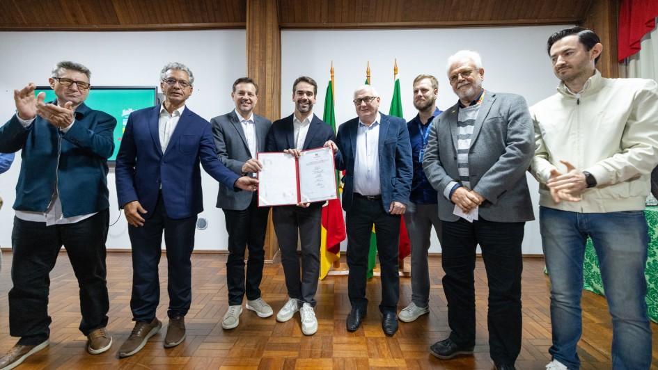 Registro coletivo em ambiente interno, com um grupo alinhado diante de bandeiras oficiais posicionadas ao fundo. No centro, um documento aberto &eacute; exibido, indicando ato formal de assinatura ou apresenta&ccedil;&atilde;o.