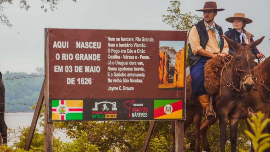 Aqui est&aacute; a descri&ccedil;&atilde;o detalhada da imagem para fins de acessibilidade:

Descri&ccedil;&atilde;o da Imagem
A fotografia mostra um marco hist&oacute;rico ao ar livre, destacando uma placa informativa e pessoas vestidas com trajes t&iacute;picos ga&uacute;chos montadas em mulas. A cena evoca a preserva&ccedil;&atilde;o da cultura e hist&oacute;ria regional.

Elementos Principais
A Placa Informativa: No centro da imagem, h&aacute; uma grande placa retangular marrom sustentada por postes de madeira.

Texto &agrave; esquerda: Em letras brancas e mai&uacute;sculas, l&ecirc;-se: "AQUI NASCEU O RIO GRANDE EM 03 DE MAIO DE 1626".

Texto ao centro: Um poema de Jayme C. Braun impresso em branco, que exalta a hist&oacute;ria do Rio Grande e de S&atilde;o Nicolau.

Lado direito da placa: Uma foto vertical de ru&iacute;nas hist&oacute;ricas (provavelmente das Miss&otilde;es).

Rodap&eacute; da placa: Exibe logotipos e bandeiras, incluindo a bandeira do Rio Grande do Sul, a bandeira de S&atilde;o Nicolau e s&iacute;mbolos de projetos tur&iacute;sticos como "Missioneiros do Pedal" e "Trilha dos Santos M&aacute;rtires".

Os Cavaleiros: &Agrave; direita da placa, um homem e uma mulher aparecem montados em mulas de pelagem escura.

Ambos vestem indument&aacute;ria tradicional ga&uacute;cha: chap&eacute;us de aba larga e copa baixa (barbicacho), len&ccedil;os no pesco&ccedil;o e coletes.

O homem, em primeiro plano, usa bombacha azul e botas de couro. A sela da mula est&aacute; coberta por um pelego de l&atilde; marrom.

O Ambiente: O cen&aacute;rio &eacute; rural, com vegeta&ccedil;&atilde;o verde baixa ao redor da placa e &aacute;rvores ao fundo. O c&eacute;u est&aacute; nublado, conferindo uma ilumina&ccedil;&atilde;o suave e difusa &agrave; imagem. &Agrave; esquerda, ao fundo, percebe-se uma extens&atilde;o de &aacute;gua (possivelmente um rio) e uma cerca de arame farpado.

Resumo visual: A imagem celebra a funda&ccedil;&atilde;o hist&oacute;rica do Rio Grande do Sul em S&atilde;o Nicolau, unindo o registro oficial do marco hist&oacute;rico &agrave; figura viva do ga&uacute;cho contempor&acirc;neo que preserva suas tradi&ccedil;&otilde;es.