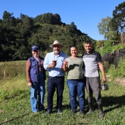 A imagem mostra um registro em &aacute;rea rural, com quatro pessoas posicionadas lado a lado em um campo gramado. Ao fundo, h&aacute; vegeta&ccedil;&atilde;o densa, &aacute;rvores altas e um relevo coberto por mata, sob c&eacute;u azul e ilumina&ccedil;&atilde;o natural intensa. Uma das pessoas segura um cart&atilde;o pl&aacute;stico identificado como Cart&atilde;o Cidad&atilde;o.