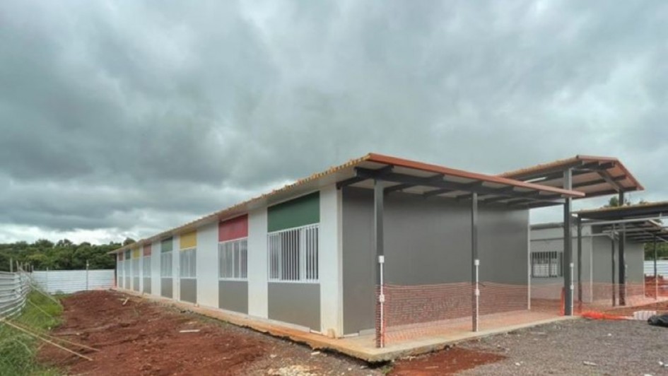 A fotografia apresenta uma constru&ccedil;&atilde;o modular t&eacute;rrea e alongada, com caracter&iacute;sticas t&iacute;picas de uma escola ou centro comunit&aacute;rio em fase final de instala&ccedil;&atilde;o. O edif&iacute;cio &eacute; composto por pain&eacute;is pr&eacute;-fabricados e est&aacute; situado em um terreno em obras.

Elementos Principais
O Edif&iacute;cio: A estrutura possui paredes brancas com detalhes coloridos (ret&acirc;ngulos em verde, vermelho e amarelo) acima das janelas, conferindo um aspecto l&uacute;dico ao projeto.

As janelas s&atilde;o brancas e possuem grades de prote&ccedil;&atilde;o verticais.

O telhado &eacute; de telhas cer&acirc;micas tradicionais (laranja), sustentado por uma estrutura met&aacute;lica preta que se estende para fora, criando uma &aacute;rea de varanda ou p&aacute;tio coberto na lateral.

O Terreno: O entorno imediato da constru&ccedil;&atilde;o mostra terra batida avermelhada e brita.

H&aacute; uma tela de prote&ccedil;&atilde;o laranja (tapume pl&aacute;stico) cercando partes da &aacute;rea externa, indicando que o local ainda &eacute; um canteiro de obras.

Ao fundo, v&ecirc;-se uma cerca de metal galvanizado e uma densa linha de &aacute;rvores verdes.

O C&eacute;u: O c&eacute;u est&aacute; densamente nublado, com nuvens carregadas em tons de cinza escuro, sugerindo um dia de chuva ou tempo inst&aacute;vel.

Resumo visual: A imagem retrata uma estrutura arquitet&ocirc;nica moderna e r&aacute;pida (modular), possivelmente destinada &agrave; educa&ccedil;&atilde;o ou atendimento p&uacute;blico, integrada a um ambiente que ainda passa por urbaniza&ccedil;&atilde;o e acabamento.