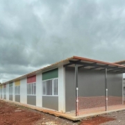 A fotografia apresenta uma constru&ccedil;&atilde;o modular t&eacute;rrea e alongada, com caracter&iacute;sticas t&iacute;picas de uma escola ou centro comunit&aacute;rio em fase final de instala&ccedil;&atilde;o. O edif&iacute;cio &eacute; composto por pain&eacute;is pr&eacute;-fabricados e est&aacute; situado em um terreno em obras.

Elementos Principais
O Edif&iacute;cio: A estrutura possui paredes brancas com detalhes coloridos (ret&acirc;ngulos em verde, vermelho e amarelo) acima das janelas, conferindo um aspecto l&uacute;dico ao projeto.

As janelas s&atilde;o brancas e possuem grades de prote&ccedil;&atilde;o verticais.

O telhado &eacute; de telhas cer&acirc;micas tradicionais (laranja), sustentado por uma estrutura met&aacute;lica preta que se estende para fora, criando uma &aacute;rea de varanda ou p&aacute;tio coberto na lateral.

O Terreno: O entorno imediato da constru&ccedil;&atilde;o mostra terra batida avermelhada e brita.

H&aacute; uma tela de prote&ccedil;&atilde;o laranja (tapume pl&aacute;stico) cercando partes da &aacute;rea externa, indicando que o local ainda &eacute; um canteiro de obras.

Ao fundo, v&ecirc;-se uma cerca de metal galvanizado e uma densa linha de &aacute;rvores verdes.

O C&eacute;u: O c&eacute;u est&aacute; densamente nublado, com nuvens carregadas em tons de cinza escuro, sugerindo um dia de chuva ou tempo inst&aacute;vel.

Resumo visual: A imagem retrata uma estrutura arquitet&ocirc;nica moderna e r&aacute;pida (modular), possivelmente destinada &agrave; educa&ccedil;&atilde;o ou atendimento p&uacute;blico, integrada a um ambiente que ainda passa por urbaniza&ccedil;&atilde;o e acabamento.