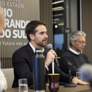 Aqui est&aacute; a descri&ccedil;&atilde;o detalhada da imagem para fins de acessibilidade:

Descri&ccedil;&atilde;o da Imagem
A fotografia registra uma coletiva de imprensa ou pronunciamento oficial. O foco principal est&aacute; no Governador do Rio Grande do Sul, Eduardo Leite, que fala ao microfone.

Elementos Principais
Eduardo Leite (Centro): O governador est&aacute; de perfil para a direita, segurando um microfone pr&oacute;ximo &agrave; boca. Ele possui barba aparada, cabelo castanho curto e veste um palet&oacute; azul-marinho sobre uma camisa branca de gola aberta. Sua express&atilde;o &eacute; s&eacute;ria e concentrada.

Outro Participante (Direita): Ao lado dele, um homem de cabelos grisalhos e &oacute;culos, vestindo um su&eacute;ter escuro sobre camisa branca, observa o evento com uma postura atenta e reservada.

Elementos Culturais sobre a Mesa: &Agrave; frente do governador, sobre uma mesa escura, destacam-se itens t&iacute;picos da cultura ga&uacute;cha: um cuia de mate com erva-verde e uma bomba de metal, al&eacute;m de uma garrafa t&eacute;rmica preta alta. Tamb&eacute;m h&aacute; copos de &aacute;gua sobre a mesa.

Plano de Fundo: Atr&aacute;s dos oradores, h&aacute; um painel decorativo com ripas verticais de madeira. No lado esquerdo do painel, l&ecirc;-se parte da identidade visual do governo: "GOVERNO DO ESTADO RIO GRANDE DO SUL" e o slogan "O futuro nos une".

Composi&ccedil;&atilde;o: A imagem utiliza uma profundidade de campo reduzida, deixando uma pessoa em primeiro plano (&agrave; esquerda, de costas) e o fundo levemente desfocados para manter o foco total nas autoridades e no pronunciamento.

Resumo visual: A imagem retrata um momento de lideran&ccedil;a e comunica&ccedil;&atilde;o governamental, contextualizado por s&iacute;mbolos da tradi&ccedil;&atilde;o ga&uacute;cha, sugerindo um an&uacute;ncio oficial ou reuni&atilde;o estrat&eacute;gica do Governo do Estado.