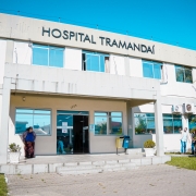 A fotografia apresenta a fachada principal do Hospital Tramanda&iacute;, capturada sob luz solar intensa e c&eacute;u azul sem nuvens. O edif&iacute;cio possui dois pavimentos e uma arquitetura funcional em tons claros.

Elementos Principais
Fachada do Edif&iacute;cio: O pr&eacute;dio &eacute; pintado de branco ou bege claro. No topo da fachada frontal, destaca-se o letreiro em letras pretas e mai&uacute;sculas: "HOSPITAL TRAMANDA&Iacute;".

Entrada: A entrada principal &eacute; protegida por uma marquise de concreto sustentada por pilares. A porta de vidro central &eacute; ladeada por grandes janelas que refletem o c&eacute;u azul. Acima da porta, v&ecirc;-se o n&uacute;mero 1255.

Pessoas e Atividade:

&Agrave; esquerda da entrada, uma mulher vestindo uma saia longa estampada e um casaco azul aguarda em p&eacute; na cal&ccedil;ada.

&Agrave; direita, no gramado, dois homens conversam; um deles veste camisa azul e cal&ccedil;a jeans, apontando em dire&ccedil;&atilde;o ao pr&eacute;dio.

Detalhes Adicionais:

Sinaliza&ccedil;&atilde;o: No canto esquerdo, em primeiro plano, h&aacute; uma placa de tr&acirc;nsito de regulamenta&ccedil;&atilde;o (proibido estacionar, com exce&ccedil;&atilde;o para embarque e desembarque r&aacute;pido).

Vegeta&ccedil;&atilde;o: O hospital &eacute; ladeado por &aacute;reas gramadas verdes, com palmeiras e arbustos decorativos em vasos brancos na entrada.

Infraestrutura: V&ecirc;-se aparelhos de ar-condicionado instalados na parede externa do segundo andar e lixeiras de coleta seletiva (azul e amarela) pr&oacute;ximas &agrave; entrada.

Resumo visual: A imagem retrata a unidade de sa&uacute;de em um dia claro, transmitindo um aspecto de organiza&ccedil;&atilde;o e atendimento p&uacute;blico regular na regi&atilde;o litor&acirc;nea.