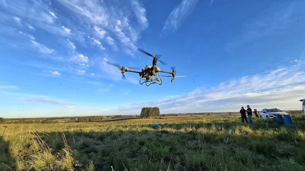 Imagem mostra drone em voo em um campo aberto, observado por tr&ecirc;s pessoas ao lado de um carro.