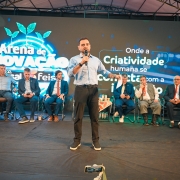 Homem discursa com microfone em um palco, atr&aacute;s dele v&aacute;rios outros homens est&atilde;o sentados e mais atr&aacute;s um grande banner com dizeres "Arena de inova&ccedil;&atilde;o"