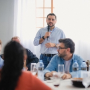 Homem fala ao microfone em um almo&ccedil;o, observado por v&aacute;rias pessoas sentadas em um ambiente de almo