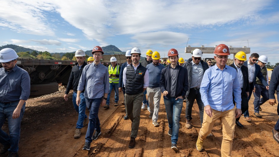 Visita t&eacute;cnica em obra de infraestrutura, com um grupo caminhando sobre solo de terra compactada. Todas as pessoas utilizam capacetes de seguran&ccedil;a e vestu&aacute;rio adequado a ambiente de obra. &Agrave; esquerda, h&aacute; caminh&otilde;es e equipamentos pesados estacionados, enquanto ao fundo surgem pilares de concreto e relevo serrano coberto por vegeta&ccedil;&atilde;o. 
