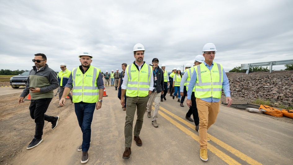 A fotografia registra uma comitiva de autoridades e t&eacute;cnicos realizando uma vistoria em uma obra rodovi&aacute;ria ao ar livre. O grupo caminha sobre uma pista de asfalto rec&eacute;m-instalada, sob um c&eacute;u nublado e cinzento.

Elementos Principais
O Grupo Central:

No centro da imagem, destaca-se o Governador Eduardo Leite, caminhando em dire&ccedil;&atilde;o &agrave; c&acirc;mera. Ele utiliza um capacete de seguran&ccedil;a branco, um colete reflexivo verde-lim&atilde;o com as inscri&ccedil;&otilde;es "Sacyr" e "Rota de Santa Maria", camisa branca e cal&ccedil;a social c&aacute;qui.

&Agrave; sua direita e esquerda, outros homens tamb&eacute;m vestem capacetes brancos e coletes reflexivos semelhantes. O homem &agrave; direita veste uma camisa azul clara e cal&ccedil;a mostarda, enquanto o da esquerda veste camisa escura e cal&ccedil;a jeans.

Composi&ccedil;&atilde;o e Movimento:

A comitiva &eacute; numerosa, com v&aacute;rias outras pessoas (homens e mulheres) caminhando logo atr&aacute;s, algumas tamb&eacute;m com equipamentos de prote&ccedil;&atilde;o individual (EPIs).

No canto esquerdo, um homem com &oacute;culos de sol e blus&atilde;o cinza caminha paralelamente ao grupo principal, segurando um celular.

O Cen&aacute;rio da Obra:

A pista possui sinaliza&ccedil;&atilde;o horizontal com duas linhas amarelas cont&iacute;nuas ao centro.

&Agrave; direita, observa-se uma encosta de pedras britadas (talude) e uma barreira met&aacute;lica de prote&ccedil;&atilde;o (guardrail) rec&eacute;m-instalada.

&Agrave; esquerda, v&ecirc;-se a lateral de uma caminhonete preta estacionada e cones de sinaliza&ccedil;&atilde;o laranjas.

O horizonte mostra uma &aacute;rea rural plana com vegeta&ccedil;&atilde;o verde ao fundo.

Resumo visual: A imagem retrata uma atividade de inspe&ccedil;&atilde;o t&eacute;cnica e oficial em uma rodovia, evidenciando o acompanhamento direto de lideran&ccedil;as governamentais em projetos de infraestrutura estadual.