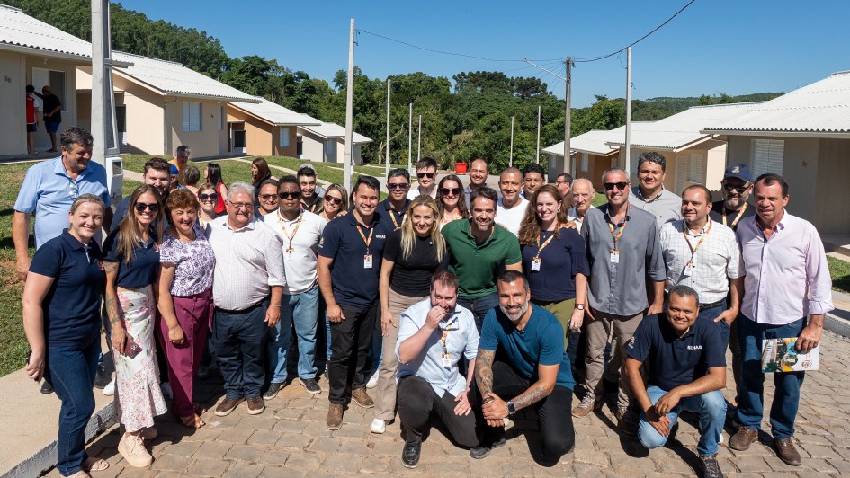 Grupo de pessoas em p&eacute; numa rua de paralelep&iacute;pedos, diante de casas novas, sob c&eacute;u azul e dia ensolarado. Muitos usam crach&aacute;s e uniformes, promovendo a participa&ccedil;&atilde;o em um projeto comunit&aacute;rio.