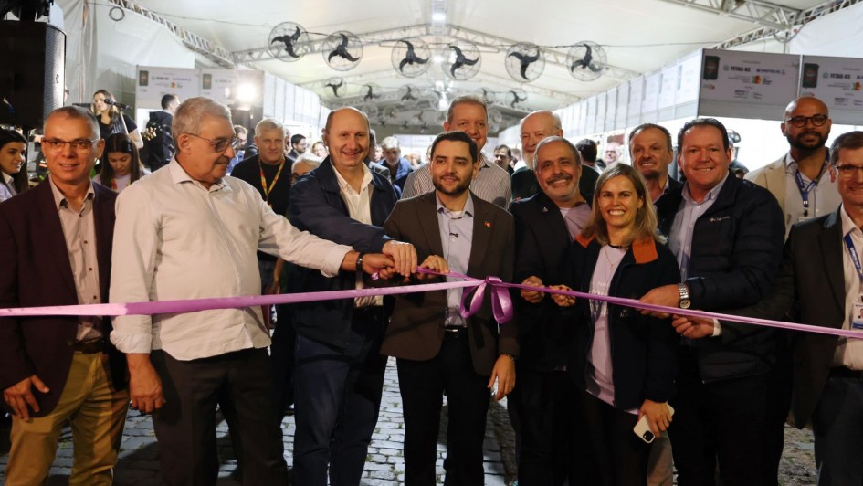 A imagem mostra um ato simb&oacute;lico de abertura realizado em um espa&ccedil;o coberto por uma grande tenda branca. No centro da cena, uma fita lil&aacute;s estendida &eacute; segurada por v&aacute;rias pessoas alinhadas, com o vice-governador ao centro do grupo. 