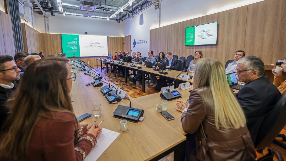 Foto mostra sala de reuni&atilde;o com diversas pessoas sentadas em torno de uma mesa no formato U, nas paredes, h&aacute; tel&otilde;es com uma apresenta&ccedil;&atilde;o de slides. 