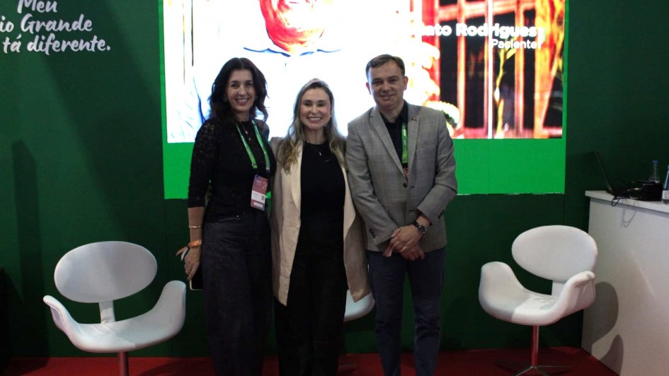 Tr&ecirc;s pessoas posam para a foto em p&eacute; em frente a um painel iluminado em um palco. 