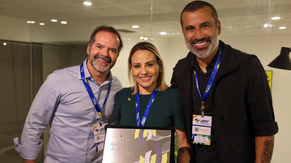 Pessoas e Elementos Centrais
O Grupo: Tr&ecirc;s pessoas est&atilde;o posicionadas lado a lado, sorrindo para a c&acirc;mera. Ao centro, uma mulher de vestido verde escuro segura uma placa de premia&ccedil;&atilde;o prateada com detalhes em amarelo e cinza, onde se l&ecirc; "Rio Grande do Sul".

Vestimenta e Identifica&ccedil;&atilde;o: &Agrave; esquerda, um homem veste uma camisa social azul clara. &Agrave; direita, um homem de barba e tatuagem no bra&ccedil;o veste uma jaqueta preta. Todos utilizam crach&aacute;s com cord&otilde;es azuis, identificando a participa&ccedil;&atilde;o no "Congresso CONSAD" (Conselho Nacional de Secret&aacute;rios de Estado da Administra&ccedil;&atilde;o).

Ambiente
Cen&aacute;rio: O local &eacute; um ambiente interno moderno, com ilumina&ccedil;&atilde;o suave e amarelada vinda de lumin&aacute;rias embutidas no teto. Ao fundo, h&aacute; divis&oacute;rias de vidro que sugerem uma sala de reuni&otilde;es ou um escrit&oacute;rio contempor&acirc;neo.

Atmosfera: A imagem transmite uma sensa&ccedil;&atilde;o de conquista, celebra&ccedil;&atilde;o e sucesso institucional.

Contexto
O t&iacute;tulo do arquivo indica que a imagem se refere ao destaque do Rio Grande do Sul em rankings de inova&ccedil;&atilde;o e efici&ecirc;ncia da m&aacute;quina p&uacute;blica.
