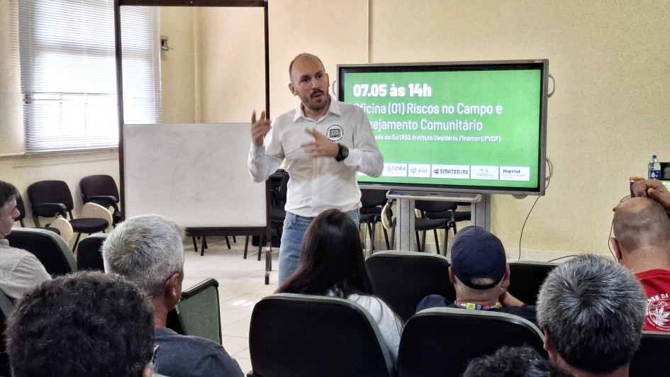 A imagem mostra uma apresenta&ccedil;&atilde;o em sala de capacita&ccedil;&atilde;o, com uma pessoa falando &agrave; frente do p&uacute;blico. Ao lado, um monitor exibe o t&iacute;tulo: &ldquo;07.05 &agrave;s 14h &ndash; Oficina (01) Riscos no Campo e Planejamento Comunit&aacute;rio&rdquo;, com logotipos institucionais ao rodap&eacute;. O p&uacute;blico permanece sentado em fileiras, voltado para a exposi&ccedil;&atilde;o. 