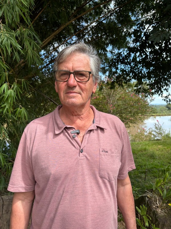 A imagem mostra um homem em área externa, posicionado em primeiro plano diante de vegetação densa com bambuzal e árvores. 
