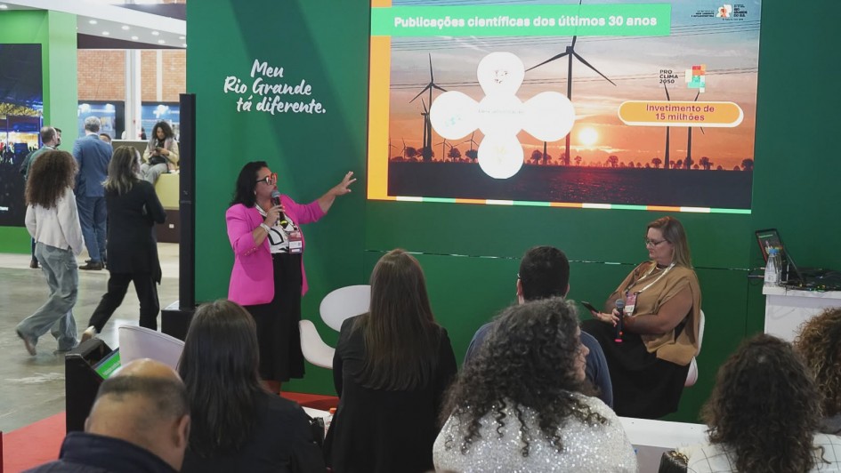 Apresenta&ccedil;&atilde;o em estande de evento, com tel&atilde;o exibindo o tema &ldquo;Publica&ccedil;&otilde;es cient&iacute;ficas dos &uacute;ltimos 30 anos&rdquo; e destaque para &ldquo;Investimento de 15 milh&otilde;es&rdquo; ligado ao programa clim&aacute;tico. A secret&aacute;ria conduz a fala com microfone enquanto o p&uacute;blico assiste sentado em &aacute;rea aberta de feira institucional.