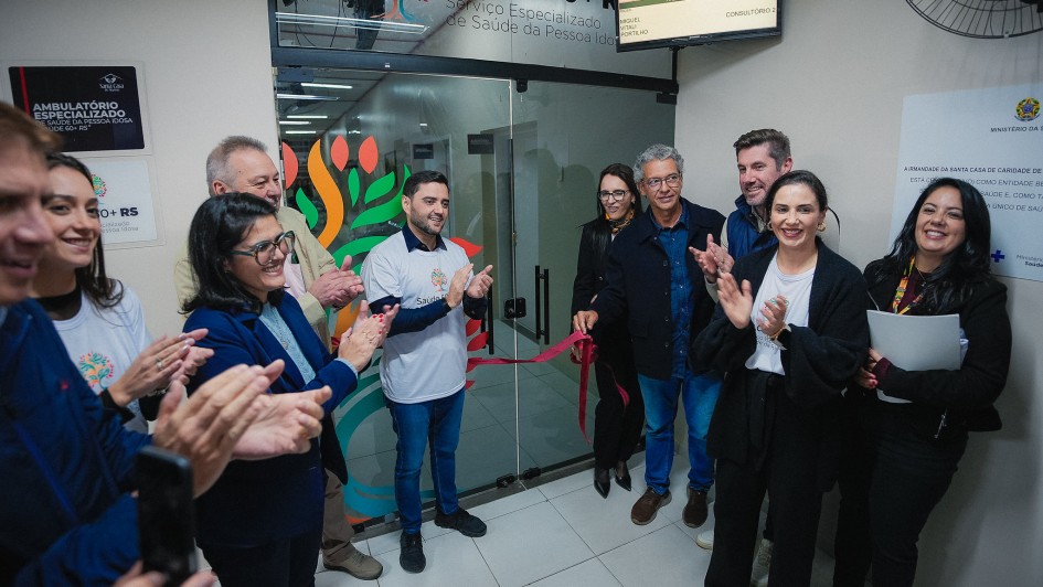 Cerim&ocirc;nia de inaugura&ccedil;&atilde;o em ambiente interno de unidade de sa&uacute;de, com grupo reunido diante de porta de vidro identificada como &ldquo;Sa&uacute;de 60+ RS &ndash; Servi&ccedil;o Especializado de Sa&uacute;de da Pessoa Idosa&rdquo;. Uma fita vermelha est&aacute; sendo cortada enquanto pessoas ao redor aplaudem.