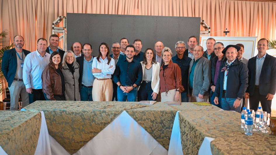Registro coletivo em ambiente interno, com grupo alinhado para foto diante de painel e estrutura de palco. Mesas com toalhas e materiais de apoio aparecem no primeiro plano.
