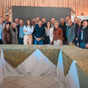 Registro coletivo em ambiente interno, com grupo alinhado para foto diante de painel e estrutura de palco. Mesas com toalhas e materiais de apoio aparecem no primeiro plano.
