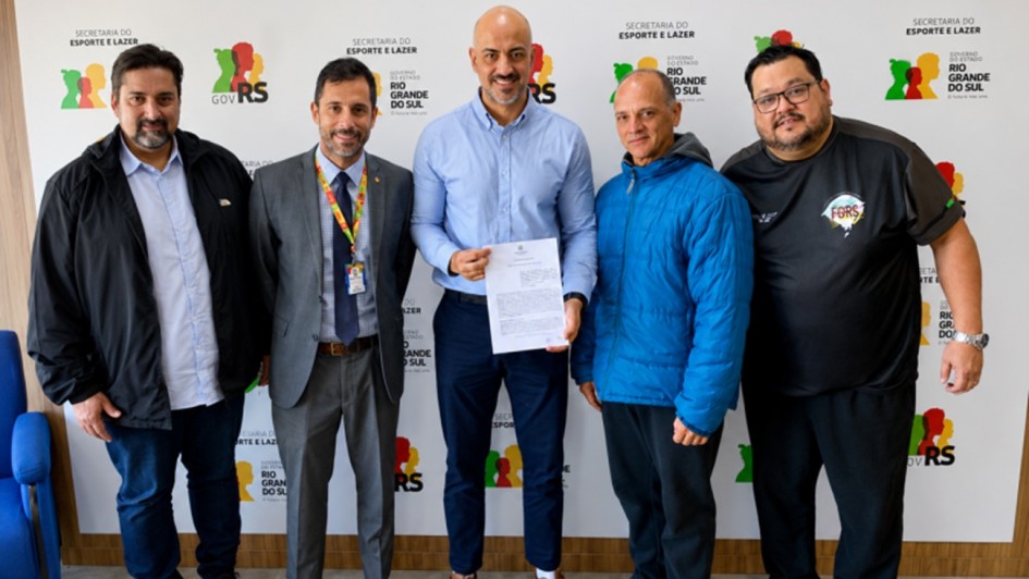 A fotografia mostra cinco homens posando para um registro oficial em um ambiente interno, posicionados &agrave; frente de um painel branco com logotipos repetidos.

Composi&ccedil;&atilde;o e vestimentas:

Ao centro: Um homem alto, branco e calvo, veste uma camisa social azul clara, cal&ccedil;as escuras e t&ecirc;nis casuais marrons. Ele segura um documento impresso com as duas m&atilde;os, apresentando-o para a foto.

&Agrave; esquerda do centro: Um homem branco, de cabelos curtos escuros, veste um terno cinza, camisa social azul, gravata escura e usa um crach&aacute; oficial com cord&atilde;o colorido.

Na extremidade esquerda: Um homem branco, com barba e cabelos escuros, veste uma jaqueta preta aberta sobre uma camisa clara e cal&ccedil;a jeans.

&Agrave; direita do centro: Um homem branco, de cabelos curtos grisalhos, veste uma jaqueta acolchoada azul vibrante e cal&ccedil;as pretas.

Na extremidade direita: Um homem branco, com &oacute;culos e barba, veste uma camiseta esportiva preta com detalhes em cinza e o logotipo "FGERS" no peito, al&eacute;m de t&ecirc;nis esportivos de cor turquesa.

Plano de fundo: O grupo est&aacute; diante de uma parede (back-drop) repleta de logotipos do Governo do Estado do Rio Grande do Sul (identificado pelo mapa estilizado em verde, vermelho e amarelo e o texto "GOV RS") e da Secretaria do Esporte e Lazer.

Ambiente: O ch&atilde;o possui um revestimento cinza claro que imita cimento queimado. &Agrave; esquerda, nota-se o bra&ccedil;o de uma poltrona azul e uma parede com acabamento em madeira.

A imagem registra a formaliza&ccedil;&atilde;o de uma parceria ou assinatura de termo entre representantes governamentais e de entidades esportivas.

