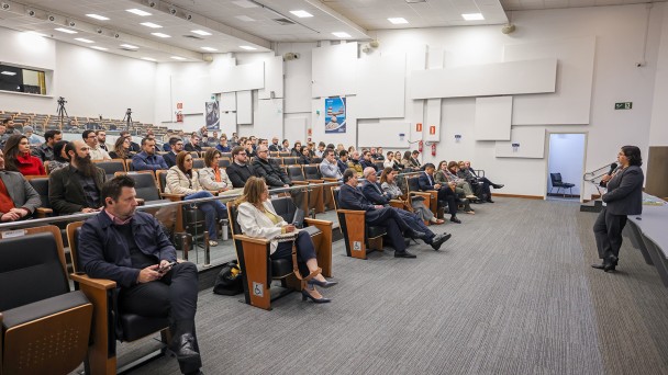 Esta &eacute; uma descri&ccedil;&atilde;o da imagem "Governo do Estado promove semin&aacute;rio com debate sobre os impactos fiscais da reforma tribut&aacute;ria.jpg" para fins de acessibilidade:

A fotografia, capturada em um plano geral e &acirc;ngulo lateral, mostra um audit&oacute;rio moderno durante a realiza&ccedil;&atilde;o de um semin&aacute;rio.

A Palestrante: &Agrave; direita da imagem, uma mulher com cabelos escuros e &oacute;culos, vestindo um conjunto formal de blazer e cal&ccedil;as cinza-escuro, fala ao microfone. Ela est&aacute; de p&eacute;, voltada para a plateia, posicionada &agrave; frente de uma mesa comprida de cor azulada que comp&otilde;e o palco.

O P&uacute;blico: O audit&oacute;rio est&aacute; ocupado por diversas pessoas sentadas em poltronas pretas com acabamento em madeira. Os participantes, homens e mulheres de v&aacute;rias idades, vestem trajes sociais e casuais, muitos deles voltados com aten&ccedil;&atilde;o para a palestrante. Algumas poltronas de ponta de fileira possuem o s&iacute;mbolo de acessibilidade para cadeirantes.

O Ambiente:

As poltronas est&atilde;o organizadas em fileiras em um plano inclinado (estilo anfiteatro).

As paredes s&atilde;o brancas e decoradas com pain&eacute;is retangulares em relevo, de diferentes tamanhos, que conferem uma textura moderna e auxiliam na ac&uacute;stica.

O teto &eacute; composto por placas modulares brancas com ilumina&ccedil;&atilde;o embutida por lumin&aacute;rias quadradas e spots direcionais.

O ch&atilde;o &eacute; revestido por um carpete cinza com listras sutis.

Ao fundo, &agrave; esquerda, &eacute; poss&iacute;vel ver um trip&eacute; com uma c&acirc;mera fotogr&aacute;fica ou de v&iacute;deo montada para registrar o evento.

&Agrave; direita, h&aacute; uma porta de sa&iacute;da aberta que revela um pequeno espa&ccedil;o interno com uma cadeira.

A imagem transmite um ambiente de di&aacute;logo t&eacute;cnico, profissionalismo e troca de conhecimentos em um evento institucional.