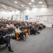 Esta &eacute; uma descri&ccedil;&atilde;o da imagem "Governo do Estado promove semin&aacute;rio com debate sobre os impactos fiscais da reforma tribut&aacute;ria.jpg" para fins de acessibilidade:

A fotografia, capturada em um plano geral e &acirc;ngulo lateral, mostra um audit&oacute;rio moderno durante a realiza&ccedil;&atilde;o de um semin&aacute;rio.

A Palestrante: &Agrave; direita da imagem, uma mulher com cabelos escuros e &oacute;culos, vestindo um conjunto formal de blazer e cal&ccedil;as cinza-escuro, fala ao microfone. Ela est&aacute; de p&eacute;, voltada para a plateia, posicionada &agrave; frente de uma mesa comprida de cor azulada que comp&otilde;e o palco.

O P&uacute;blico: O audit&oacute;rio est&aacute; ocupado por diversas pessoas sentadas em poltronas pretas com acabamento em madeira. Os participantes, homens e mulheres de v&aacute;rias idades, vestem trajes sociais e casuais, muitos deles voltados com aten&ccedil;&atilde;o para a palestrante. Algumas poltronas de ponta de fileira possuem o s&iacute;mbolo de acessibilidade para cadeirantes.

O Ambiente:

As poltronas est&atilde;o organizadas em fileiras em um plano inclinado (estilo anfiteatro).

As paredes s&atilde;o brancas e decoradas com pain&eacute;is retangulares em relevo, de diferentes tamanhos, que conferem uma textura moderna e auxiliam na ac&uacute;stica.

O teto &eacute; composto por placas modulares brancas com ilumina&ccedil;&atilde;o embutida por lumin&aacute;rias quadradas e spots direcionais.

O ch&atilde;o &eacute; revestido por um carpete cinza com listras sutis.

Ao fundo, &agrave; esquerda, &eacute; poss&iacute;vel ver um trip&eacute; com uma c&acirc;mera fotogr&aacute;fica ou de v&iacute;deo montada para registrar o evento.

&Agrave; direita, h&aacute; uma porta de sa&iacute;da aberta que revela um pequeno espa&ccedil;o interno com uma cadeira.

A imagem transmite um ambiente de di&aacute;logo t&eacute;cnico, profissionalismo e troca de conhecimentos em um evento institucional.