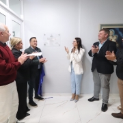 A imagem mostra um ato de inaugura&ccedil;&atilde;o em ambiente interno, com grupo reunido diante de uma placa de identifica&ccedil;&atilde;o fixada na parede, sendo revelada com faixa. As pessoas est&atilde;o dispostas ao redor e aplaudem ap&oacute;s a cerim&ocirc;nia.