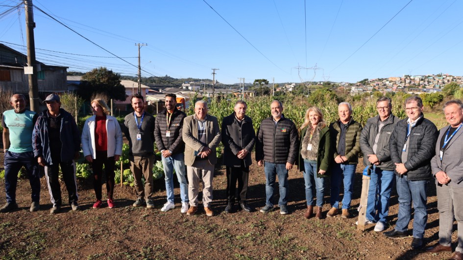 Registro coletivo em horta comunit&aacute;ria, com grupo alinhado em &aacute;rea de cultivo ao ar livre. Ao fundo, h&aacute; canteiros com plantas, casas e rede el&eacute;trica.