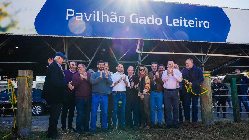 Registro coletivo em frente ao Pavilh&atilde;o Gado Leiteiro, durante ato oficial com corte simb&oacute;lico de fita nas laterais. Um grupo est&aacute; alinhado na entrada, com estrutura met&aacute;lica do galp&atilde;o ao fundo e luz do sol incidindo sobre a cena, marcando a abertura do espa&ccedil;o expositivo.