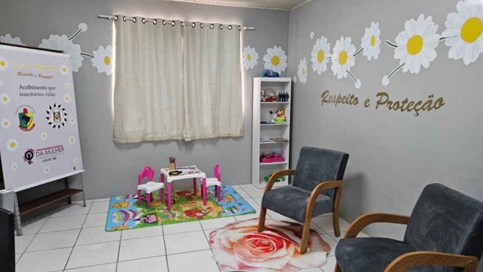 Esta &eacute; uma descri&ccedil;&atilde;o da imagem "Pol&iacute;cia Civil inaugura Sala das Margaridas em Quara&iacute;.jpeg" para fins de acessibilidade:

A fotografia mostra o interior de uma sala de acolhimento humanizado, decorada em tons de cinza, branco e rosa, projetada para oferecer conforto e seguran&ccedil;a.

Mobili&aacute;rio de Acolhimento: No centro e &agrave; direita da imagem, h&aacute; duas poltronas de estofado cinza escuro com bra&ccedil;os de madeira clara, posicionadas sobre um tapete circular que estampa uma grande rosa em tons de rosa e branco.

Espa&ccedil;o Infantil: &Agrave; esquerda, sobre um tapete colorido com desenhos l&uacute;dicos, encontra-se um conjunto de mesa branca com pernas rosa e duas cadeiras pequenas (uma rosa e uma branca), destinado a crian&ccedil;as.

Decora&ccedil;&atilde;o das Paredes:

As paredes cinzas s&atilde;o decoradas com adesivos de grandes margaridas brancas com miolo amarelo, conectadas por tra&ccedil;os que simulam ramos.

Na parede &agrave; direita, l&ecirc;-se a frase "Respeito e Prote&ccedil;&atilde;o" escrita em letras douradas com relevo.

Ao fundo, uma janela &eacute; coberta por cortinas claras de tecido leve.

Elementos de Apoio:

Entre a janela e as poltronas, h&aacute; uma estante branca estreita com prateleiras contendo brinquedos e objetos decorativos.

No canto esquerdo, um banner vertical branco exibe o nome "Sala das Margaridas", os logotipos da Pol&iacute;cia Civil e do Governo do Estado, al&eacute;m da frase "Acolhimento que transforma vidas" e informa&ccedil;&otilde;es da Procuradoria Especial da Mulher.

A imagem transmite uma atmosfera de tranquilidade e cuidado, caracter&iacute;stica dos espa&ccedil;os voltados ao atendimento de mulheres e crian&ccedil;as em situa&ccedil;&atilde;o de vulnerabilidade.
