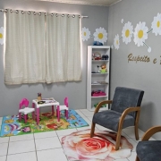 Esta &eacute; uma descri&ccedil;&atilde;o da imagem "Pol&iacute;cia Civil inaugura Sala das Margaridas em Quara&iacute;.jpeg" para fins de acessibilidade:

A fotografia mostra o interior de uma sala de acolhimento humanizado, decorada em tons de cinza, branco e rosa, projetada para oferecer conforto e seguran&ccedil;a.

Mobili&aacute;rio de Acolhimento: No centro e &agrave; direita da imagem, h&aacute; duas poltronas de estofado cinza escuro com bra&ccedil;os de madeira clara, posicionadas sobre um tapete circular que estampa uma grande rosa em tons de rosa e branco.

Espa&ccedil;o Infantil: &Agrave; esquerda, sobre um tapete colorido com desenhos l&uacute;dicos, encontra-se um conjunto de mesa branca com pernas rosa e duas cadeiras pequenas (uma rosa e uma branca), destinado a crian&ccedil;as.

Decora&ccedil;&atilde;o das Paredes:

As paredes cinzas s&atilde;o decoradas com adesivos de grandes margaridas brancas com miolo amarelo, conectadas por tra&ccedil;os que simulam ramos.

Na parede &agrave; direita, l&ecirc;-se a frase "Respeito e Prote&ccedil;&atilde;o" escrita em letras douradas com relevo.

Ao fundo, uma janela &eacute; coberta por cortinas claras de tecido leve.

Elementos de Apoio:

Entre a janela e as poltronas, h&aacute; uma estante branca estreita com prateleiras contendo brinquedos e objetos decorativos.

No canto esquerdo, um banner vertical branco exibe o nome "Sala das Margaridas", os logotipos da Pol&iacute;cia Civil e do Governo do Estado, al&eacute;m da frase "Acolhimento que transforma vidas" e informa&ccedil;&otilde;es da Procuradoria Especial da Mulher.

A imagem transmite uma atmosfera de tranquilidade e cuidado, caracter&iacute;stica dos espa&ccedil;os voltados ao atendimento de mulheres e crian&ccedil;as em situa&ccedil;&atilde;o de vulnerabilidade.