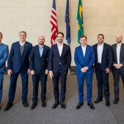 A fotografia mostra sete homens posando em p&eacute;, lado a lado, em um ambiente interno amplo com est&eacute;tica institucional.

O Grupo:

No centro da imagem est&aacute; o governador Eduardo Leite, um homem branco de barba e cabelos castanhos curtos, vestindo um terno azul-marinho sobre camisa branca sem gravata.

Ele est&aacute; ladeado por outros seis homens, todos brancos e vestindo trajes sociais (ternos em tons de azul e cinza-escuro com camisas claras).

Todos os integrantes do grupo sorriem para a c&acirc;mera e mant&ecirc;m uma postura formal, a maioria com as m&atilde;os cruzadas &agrave; frente do corpo.

Plano de Fundo:

Logo atr&aacute;s de Eduardo Leite, encontram-se tr&ecirc;s bandeiras em seus respectivos mastros: a bandeira dos Estados Unidos (&agrave; esquerda), uma bandeira azul (ao centro) e a bandeira do Brasil (&agrave; direita).

A parede ao fundo &eacute; composta por grandes blocos de pedra clara ou cer&acirc;mica, conferindo um aspecto s&oacute;brio e moderno ao local.

O Ambiente:

O ch&atilde;o &eacute; revestido por um carpete cinza escuro uniforme.

A ilumina&ccedil;&atilde;o &eacute; clara e distribu&iacute;da por igual em toda a cena.

A imagem registra um encontro oficial de natureza diplom&aacute;tica ou de neg&oacute;cios entre representantes governamentais e investidores.