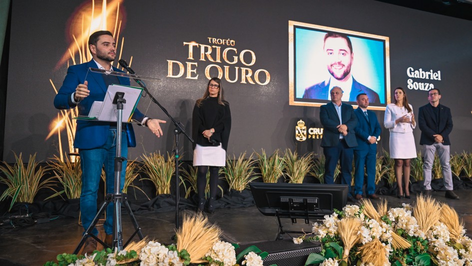 Vista ampliada do palco durante cerim&ocirc;nia do Trof&eacute;u Trigo de Ouro, com grupo alinhado ao lado da &aacute;rea de fala, onde o vice-governador est&aacute; posicionado ao microfone. Ao fundo, tel&atilde;o apresenta a identifica&ccedil;&atilde;o &ldquo;Gabriel Souza&rdquo; e a identidade visual do evento, com decora&ccedil;&atilde;o tem&aacute;tica com plantas e arranjos.