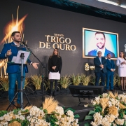 Vista ampliada do palco durante cerim&ocirc;nia do Trof&eacute;u Trigo de Ouro, com grupo alinhado ao lado da &aacute;rea de fala, onde o vice-governador est&aacute; posicionado ao microfone. Ao fundo, tel&atilde;o apresenta a identifica&ccedil;&atilde;o &ldquo;Gabriel Souza&rdquo; e a identidade visual do evento, com decora&ccedil;&atilde;o tem&aacute;tica com plantas e arranjos.