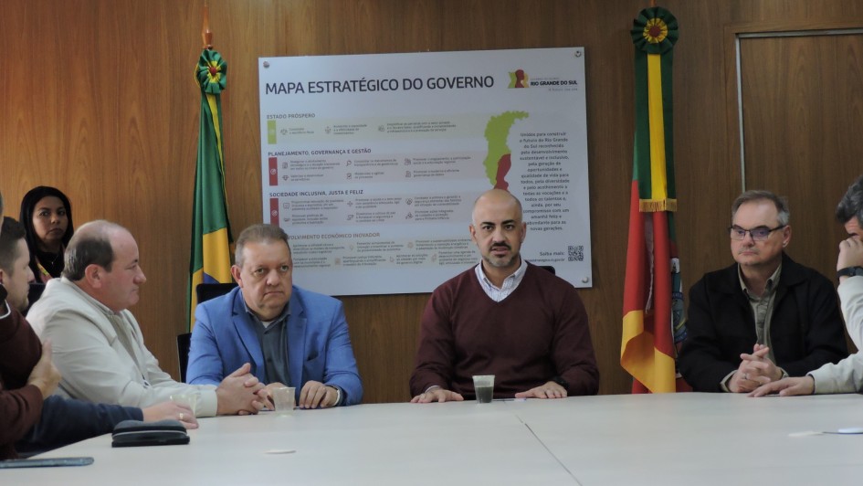 A fotografia mostra uma reuni&atilde;o oficial em torno de uma mesa branca, com foco em quatro homens sentados ao centro e &agrave; direita.

Os Participantes:

Ao centro, um homem branco e calvo, vestindo um su&eacute;ter bord&ocirc; sobre camisa clara, fala para o grupo. Ele tem um copo pequeno &agrave; sua frente.

&Agrave; sua esquerda (centro-esquerda da foto), um homem branco veste um palet&oacute; azul sobre camisa clara e observa atentamente.

Na extremidade esquerda, um homem de palet&oacute; bege est&aacute; de perfil, com as m&atilde;os entrela&ccedil;adas sobre a mesa.

&Agrave; direita do homem de bord&ocirc;, um homem de &oacute;culos e casaco preto tamb&eacute;m acompanha a fala com as m&atilde;os sobre a mesa.

No canto superior esquerdo, o rosto de uma mulher &eacute; vis&iacute;vel ao fundo.

Plano de Fundo:

Atr&aacute;s do grupo, h&aacute; um grande painel branco intitulado "MAPA ESTRAT&Eacute;GICO DO GOVERNO", contendo textos, &iacute;cones coloridos e o logotipo do Governo do Estado do Rio Grande do Sul.

Ladeando o painel, est&atilde;o duas bandeiras em seus mastros: a do Brasil &agrave; esquerda e a do Rio Grande do Sul &agrave; direita.

O Ambiente:

A parede &eacute; revestida com pain&eacute;is de madeira em tom m&eacute;dio.

A ilumina&ccedil;&atilde;o &eacute; frontal e clara, vinda de cima.

Sobre a mesa branca e limpa, veem-se apenas alguns copos descart&aacute;veis e um estojo de &oacute;culos preto.

A imagem registra um momento de coordena&ccedil;&atilde;o pol&iacute;tica e administrativa, focada no planejamento estrat&eacute;gico do estado.