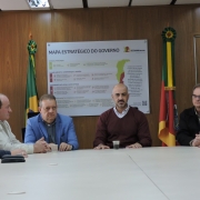 A fotografia mostra uma reuni&atilde;o oficial em torno de uma mesa branca, com foco em quatro homens sentados ao centro e &agrave; direita.

Os Participantes:

Ao centro, um homem branco e calvo, vestindo um su&eacute;ter bord&ocirc; sobre camisa clara, fala para o grupo. Ele tem um copo pequeno &agrave; sua frente.

&Agrave; sua esquerda (centro-esquerda da foto), um homem branco veste um palet&oacute; azul sobre camisa clara e observa atentamente.

Na extremidade esquerda, um homem de palet&oacute; bege est&aacute; de perfil, com as m&atilde;os entrela&ccedil;adas sobre a mesa.

&Agrave; direita do homem de bord&ocirc;, um homem de &oacute;culos e casaco preto tamb&eacute;m acompanha a fala com as m&atilde;os sobre a mesa.

No canto superior esquerdo, o rosto de uma mulher &eacute; vis&iacute;vel ao fundo.

Plano de Fundo:

Atr&aacute;s do grupo, h&aacute; um grande painel branco intitulado "MAPA ESTRAT&Eacute;GICO DO GOVERNO", contendo textos, &iacute;cones coloridos e o logotipo do Governo do Estado do Rio Grande do Sul.

Ladeando o painel, est&atilde;o duas bandeiras em seus mastros: a do Brasil &agrave; esquerda e a do Rio Grande do Sul &agrave; direita.

O Ambiente:

A parede &eacute; revestida com pain&eacute;is de madeira em tom m&eacute;dio.

A ilumina&ccedil;&atilde;o &eacute; frontal e clara, vinda de cima.

Sobre a mesa branca e limpa, veem-se apenas alguns copos descart&aacute;veis e um estojo de &oacute;culos preto.

A imagem registra um momento de coordena&ccedil;&atilde;o pol&iacute;tica e administrativa, focada no planejamento estrat&eacute;gico do estado.