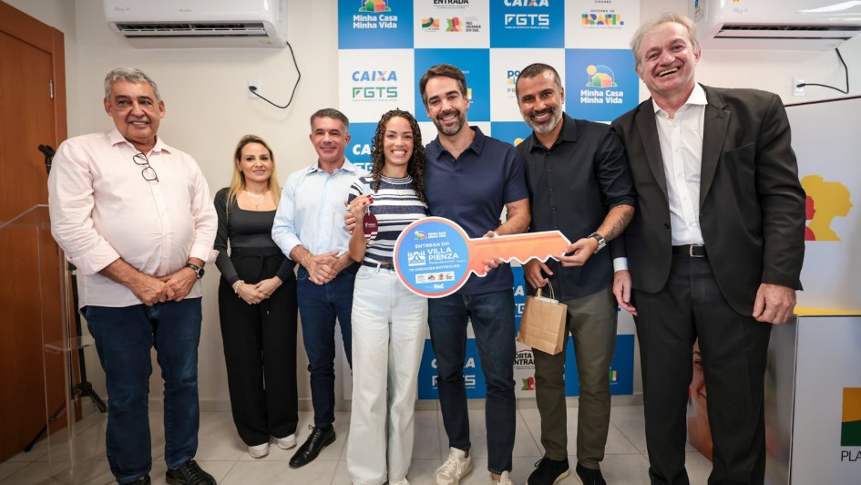 A fotografia mostra um grupo de sete pessoas sorridentes em um ato oficial de entrega de chaves, em um ambiente interno.

A&ccedil;&atilde;o Central: No centro, o governador Eduardo Leite e um homem &agrave; sua direita seguram juntos uma chave simb&oacute;lica gigante feita de material r&iacute;gido. A chave &eacute; colorida (azul, laranja e branco) e cont&eacute;m o texto "ENTREGA DO VILLA PIENZA RESIDENCIAL". Entre eles, uma mulher jovem, de cabelos cacheados, segura um molho de chaves real com um chaveiro vermelho, representando a benefici&aacute;ria do programa.

Os Participantes:

Eduardo Leite (ao centro) veste uma camisa polo azul-marinho, cal&ccedil;a jeans escura e t&ecirc;nis claros.

&Agrave; esquerda da benefici&aacute;ria, tr&ecirc;s pessoas observam: um homem de camisa social azul clara, uma mulher de blusa preta e um homem de camisa rosa clara.

&Agrave; direita do grupo central, al&eacute;m do homem que segura a chave gigante (de camisa preta e bra&ccedil;o tatuado), h&aacute; um homem de terno preto e cabelos grisalhos que sorri abertamente para a c&acirc;mera.

Plano de Fundo: Atr&aacute;s do grupo, h&aacute; um painel (back-drop) com um padr&atilde;o de quadrados azuis e brancos contendo logotipos oficiais: "Minha Casa Minha Vida", "Porta de Entrada", "Caixa FGTS" e o logotipo do Governo Federal (Brasil).

O Ambiente: A sala tem paredes brancas e um aparelho de ar-condicionado do tipo split instalado no alto, &agrave; esquerda. O ch&atilde;o &eacute; de piso cer&acirc;mico claro.

A imagem registra um momento de celebra&ccedil;&atilde;o pela entrega de moradias populares e parcerias habitacionais.