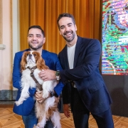 Registro institucional em ambiente interno, com duas pessoas posicionadas lado a lado enquanto uma delas segura um cachorro de pequeno porte com pelagem branca e marrom. Ao fundo, h&aacute; um painel digital colorido com elementos gr&aacute;ficos e texto relacionado a fundo de prote&ccedil;&atilde;o e bem-estar animal.