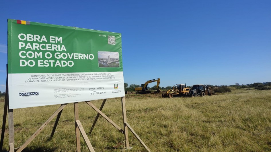 Foto mostra campo onde est&aacute; sendo feita a obra. Ao fundo m&aacute;quinas trabalham no terreno. Em primeiro plano, uma placa do governo do Estado sobre a obra.